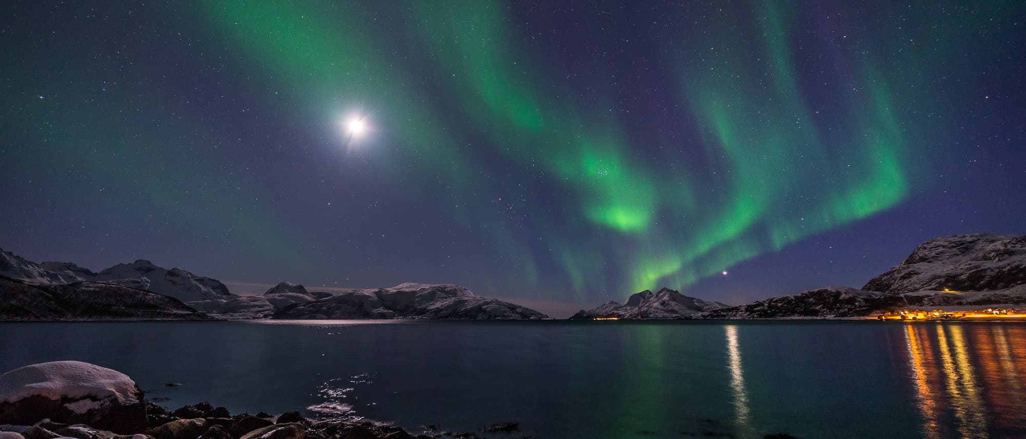 Ein n&auml;chtliches Foto zeigt das Nordlicht, das in gr&uuml;nen und violetten Wellen &uuml;ber einem klaren Sternenhimmel tanzt. Im Vordergrund sind schneebedeckte Felsen am Ufer eines ruhigen Sees zu sehen, der die Lichter reflektiert. Am Horizont sind schneebedeckte Berge und ein kleiner beleuchteter Ort zu erkennen. Der Mond leuchtet hell am Himmel.