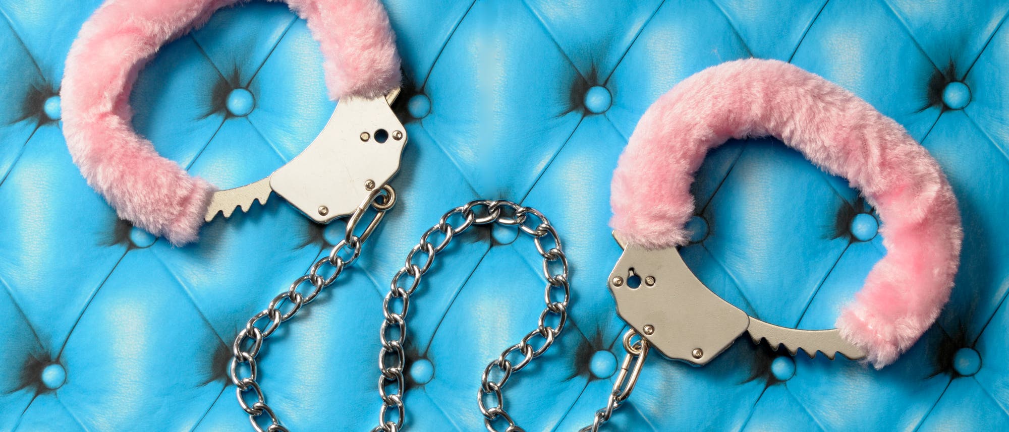 Ein Paar Handschellen mit rosa Plüschüberzug liegt auf einer blauen, gesteppten Oberfläche. Die Handschellen sind durch eine silberne Kette verbunden. Die Szene vermittelt einen Kontrast zwischen der weichen Textur des Plüschs und der metallischen Härte der Handschellen.