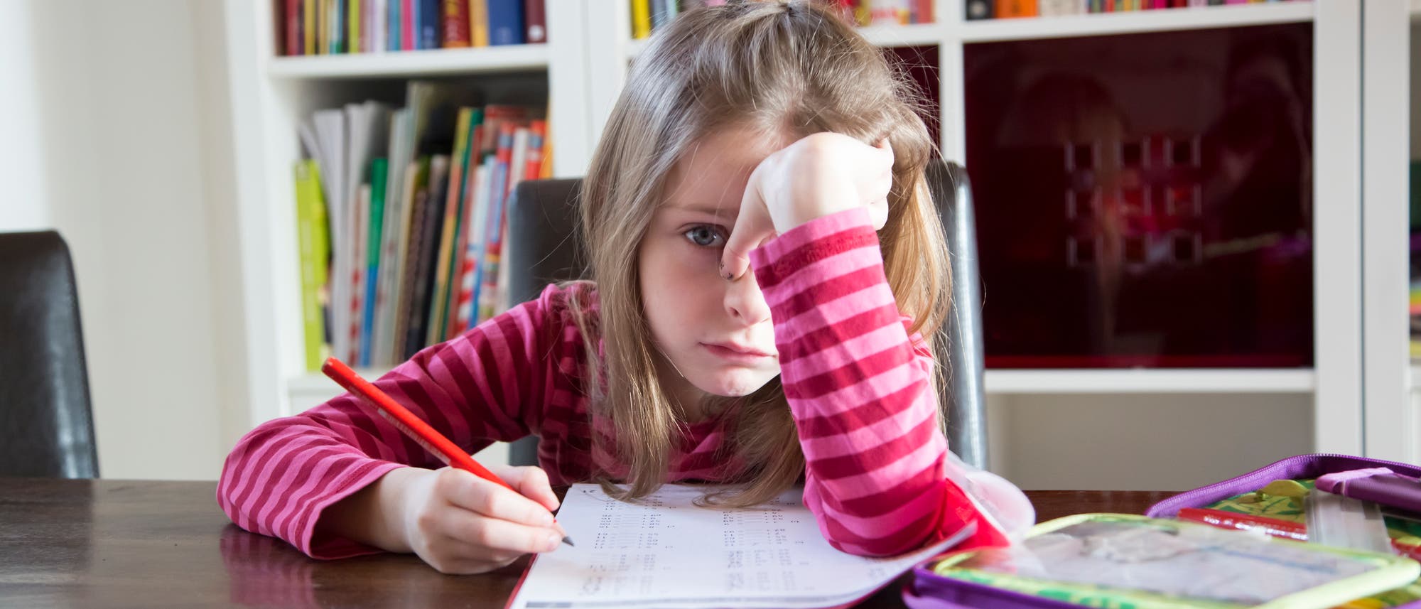 Ein M&auml;dchen im Grundschulalter sitzt an einem Tisch und macht Hausaufgaben. Es h&auml;lt einen Stift in der Hand, st&uuml;tzt den Kopf auf die andere Hand und guckt traurig. Im Hintergrund ist ein B&uuml;cherregal mit bunten B&uuml;chern zu sehen. Auf dem Tisch liegt ein offenes Federm&auml;ppchen.