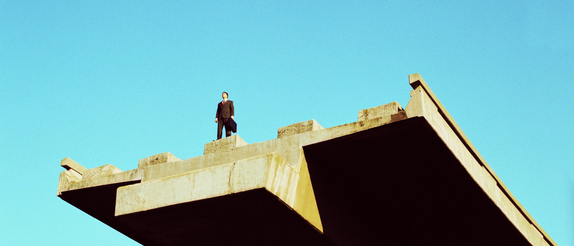 Eine Person in dunklem Mantel steht an der Kante eines unvollendeten Brückenabschnitts. Der Himmel ist klar und blau. Die Brücke aus Beton endet abrupt, was den Eindruck von Unvollständigkeit und offener Zukunft vermittelt.