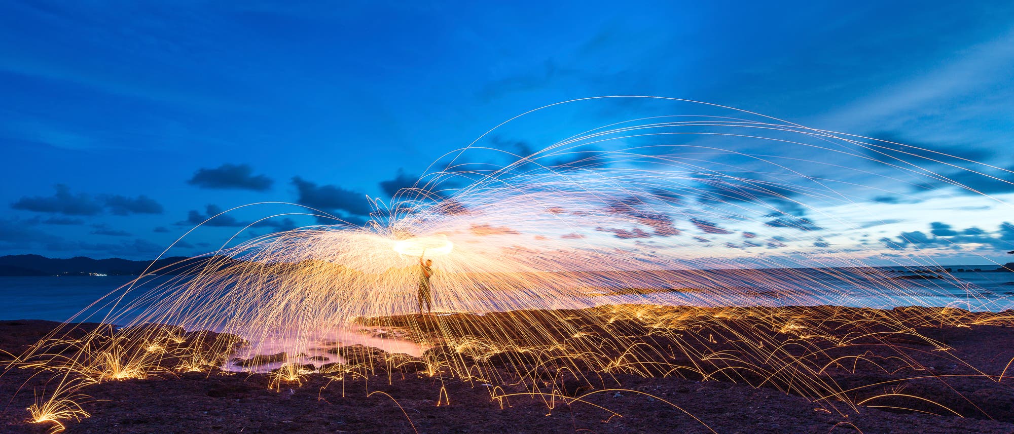 Eine Person steht auf einem felsigen Strand bei Sonnenuntergang und schwingt brennende Stahlwolle, wodurch Funken in einem weiten Bogen sprühen. Der Himmel ist in tiefem Blau mit vereinzelten Wolken gefärbt, und das Meer ist im Hintergrund sichtbar. Die Funken erzeugen leuchtende, geschwungene Linien, die sich über den Boden erstrecken.