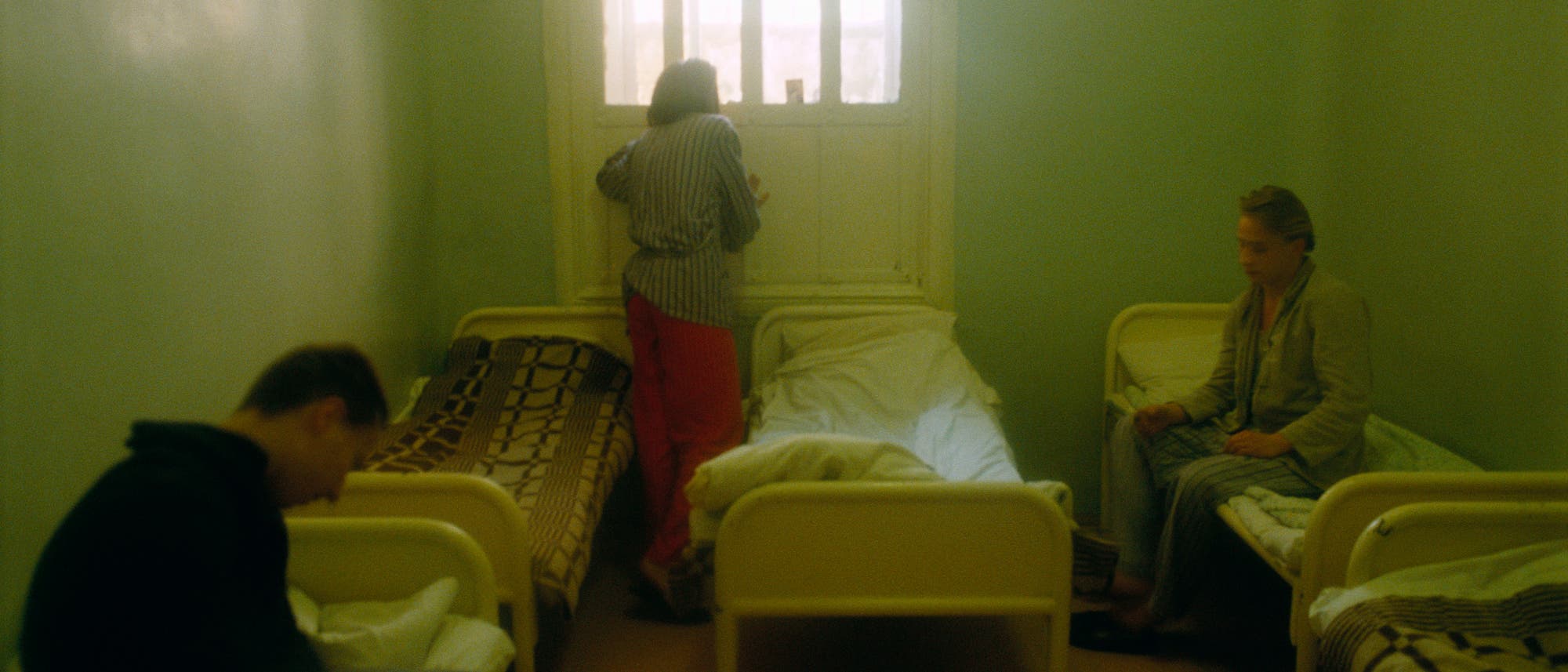 Ein karger Raum mit drei Betten, in dem drei Personen zu sehen sind. Eine Person sitzt auf einem Bett links im Vordergrund, eine andere steht am Fenster in der Mitte des Raumes, und die dritte sitzt auf einem Bett rechts. Der Raum ist in ged&auml;mpften Farben gehalten, mit blassen W&auml;nden und einem Fenster, das diffuses Licht hereinl&auml;sst. Die Atmosph&auml;re wirkt beengt und nachdenklich.