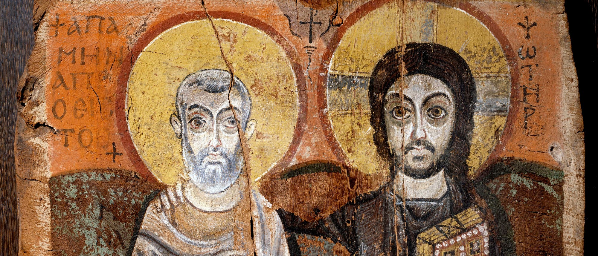 Ein antikes Gemälde auf Holz zeigt zwei heilige Figuren mit Heiligenscheinen. Die linke Figur trägt ein langes Gewand und hält eine Schriftrolle, während die rechte Figur ein reichverziertes Buch hält. Beide Figuren sind frontal dargestellt und von griechischem Text umgeben. Der Hintergrund ist in warmen Erdtönen gehalten, was dem Bild eine historische und religiöse Atmosphäre verleiht.
