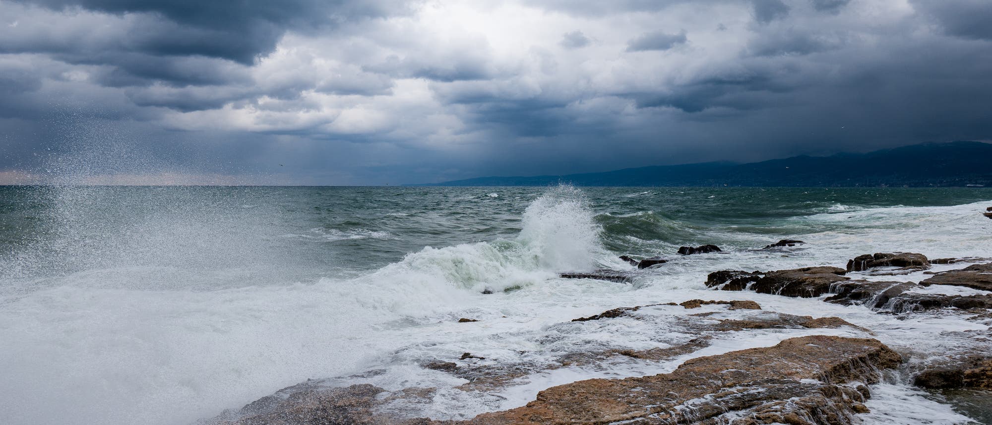 St&uuml;rmische Meeresk&uuml;ste mit dunklen Wolken am Himmel. Wellen schlagen gegen die Felsen, w&auml;hrend Gischt in die Luft spritzt. Die Szene vermittelt eine dynamische und dramatische Atmosph&auml;re. Im Hintergrund sind entfernte H&uuml;gel zu erkennen.