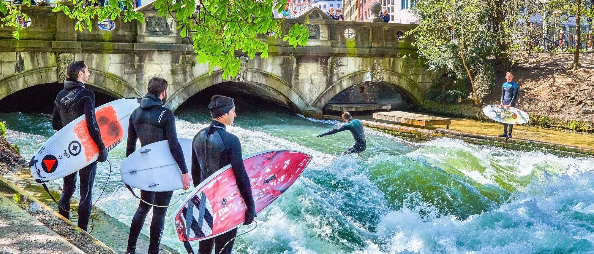 Surfer in Neoprenanzügen stehen mit ihren Surfbrettern am Ufer eines Flusses, während ein Surfer auf einer stehenden Welle reitet. Im Hintergrund ist eine Steinbrücke mit Bögen zu sehen, und Zuschauer beobachten das Geschehen. Die Szene spielt sich in einer urbanen Umgebung mit Gebäuden und Bäumen ab.