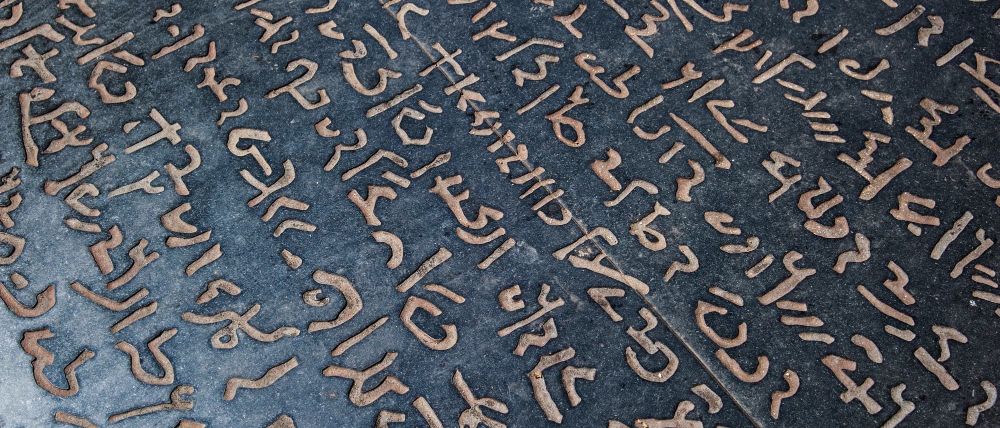Eine Steinplatte mit eingravierten Schriftzeichen, die in mehreren Reihen angeordnet sind. Die Zeichen sind in einer antiken oder unbekannten Schriftart gestaltet und heben sich durch ihre helle Farbe vom dunklen Hintergrund ab. Die Anordnung der Zeichen wirkt geordnet und systematisch, was auf eine historische oder kulturelle Bedeutung hinweisen könnte. Eine Steinplatte mit eingravierten Schriftzeichen, die in mehreren Reihen angeordnet sind. Die Zeichen sind in einer antiken oder unbekannten Schriftart gestaltet und heben sich durch ihre helle Farbe vom dunklen Hintergrund ab. Die Anordnung der Zeichen wirkt geordnet und systematisch, was auf eine historische oder kulturelle Bedeutung hinweisen könnte.