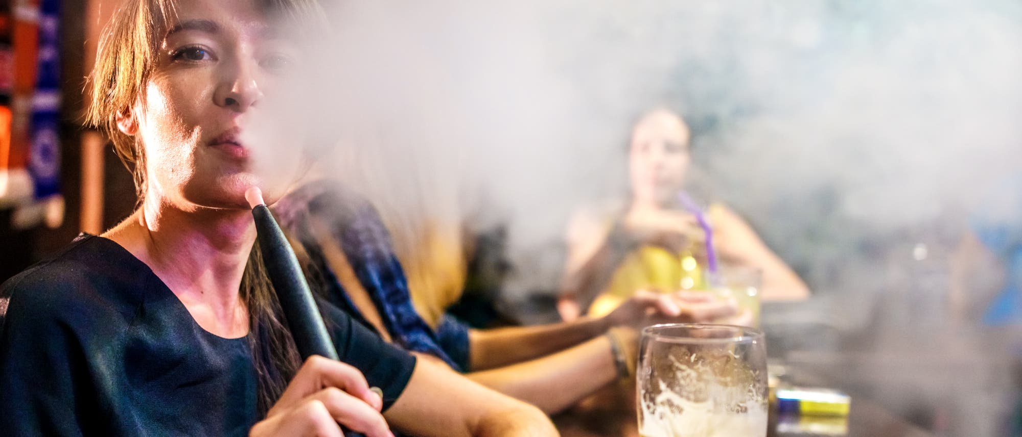 Eine Person sitzt in einer Bar und raucht eine Shisha, wobei viel Rauch die Umgebung f&uuml;llt. Im Vordergrund steht ein Glas mit einem dunklen Getr&auml;nk auf dem Tisch. Im Hintergrund sind weitere Personen zu sehen, die ebenfalls Shisha rauchen. Die Szene vermittelt eine entspannte, gesellige Atmosph&auml;re.