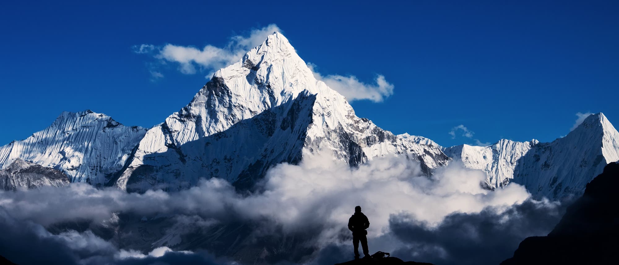 Der Mount Everest ragt weißgrau und schroff in den blauen Himmel auf, eine kleine weiße Wolke befindet sich hinter seinem Gipfel. Im Vordergrund schweben ebenfalls kleinere weiße Wolken in tieferen Lagen. Die schwarze Silhouette eines Mannes steht auf einem schwaren Felsen im Vordergrund