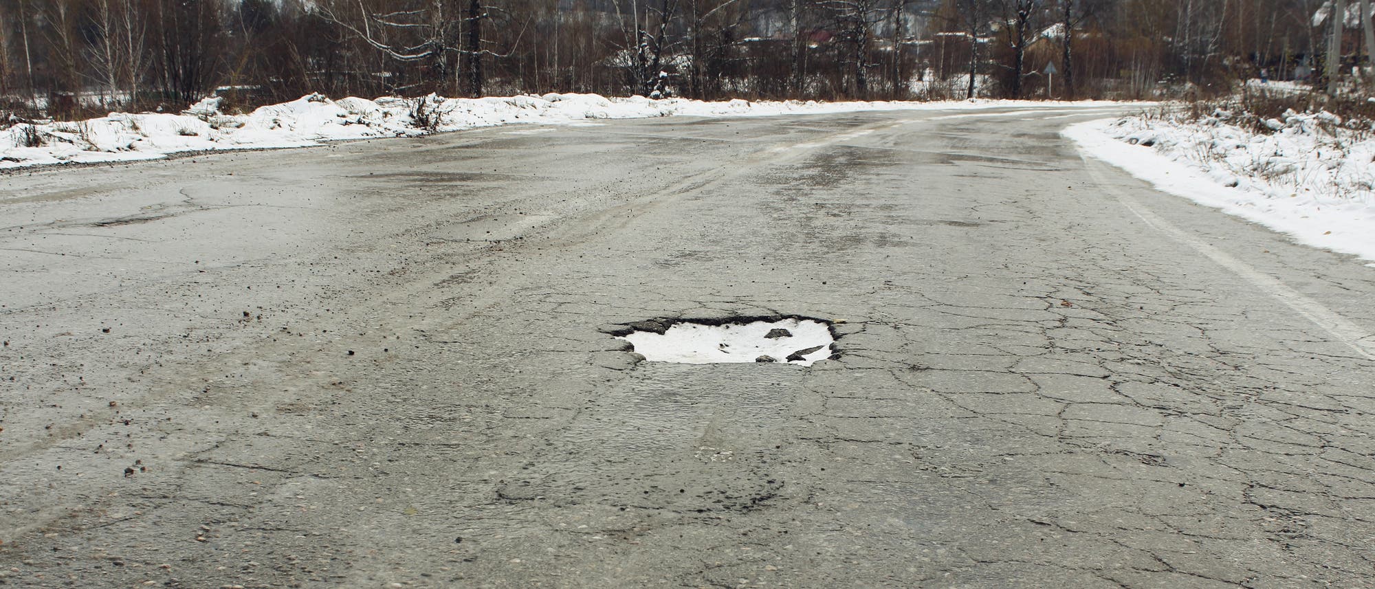 Im rissigen Asphalt einer Straße im Winter klafft ein großes, mit verfestigtem Schnee gefülltes Schlagloch