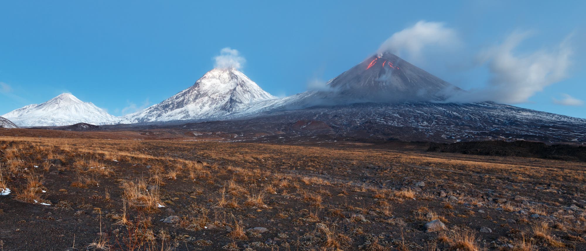 Drei schneebedeckte Vulkane unter klarem Himmel. Der rechte Vulkan zeigt Anzeichen von Aktivität mit aufsteigendem Rauch und glühender Lava an der Spitze. Im Vordergrund eine karge, mit Gras bewachsene Landschaft.