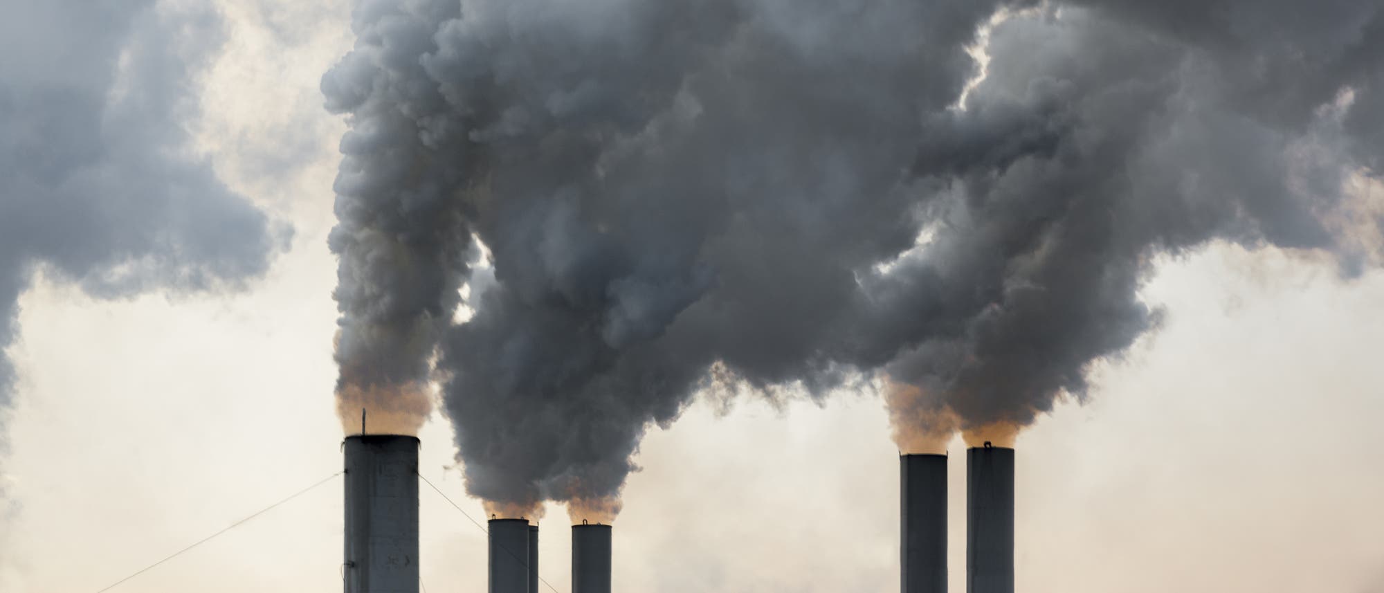 Rauch steigt aus mehreren Schornsteinen eines Kraftwerks in den Himmel. Die dichten, dunklen Rauchwolken kontrastieren mit dem helleren Hintergrund des Himmels. Das Bild thematisiert Umweltverschmutzung und industrielle Emissionen.