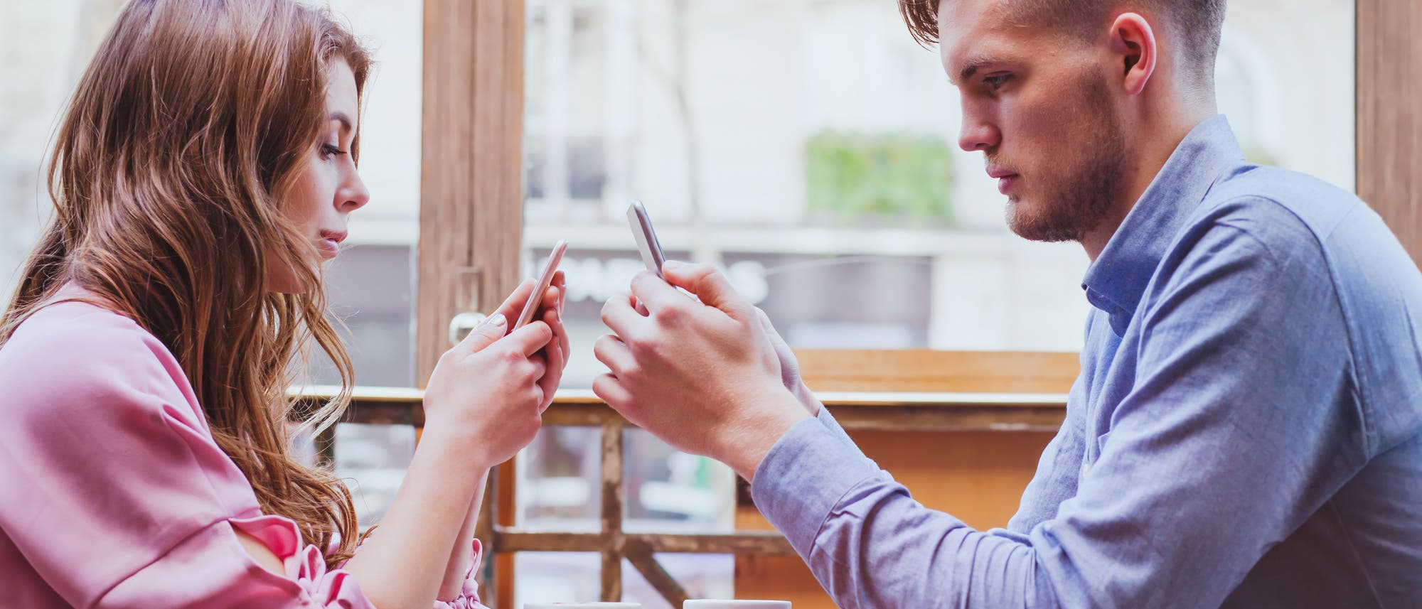 Zwei Personen sitzen sich in einem Caf&eacute; gegen&uuml;ber und schauen auf ihre Smartphones. Auf dem Tisch stehen zwei leere Tassen. Die Szene vermittelt den Eindruck von moderner Kommunikation und Technologie im Alltag. Im Hintergrund sind Fenster mit Blick auf eine st&auml;dtische Umgebung zu sehen.