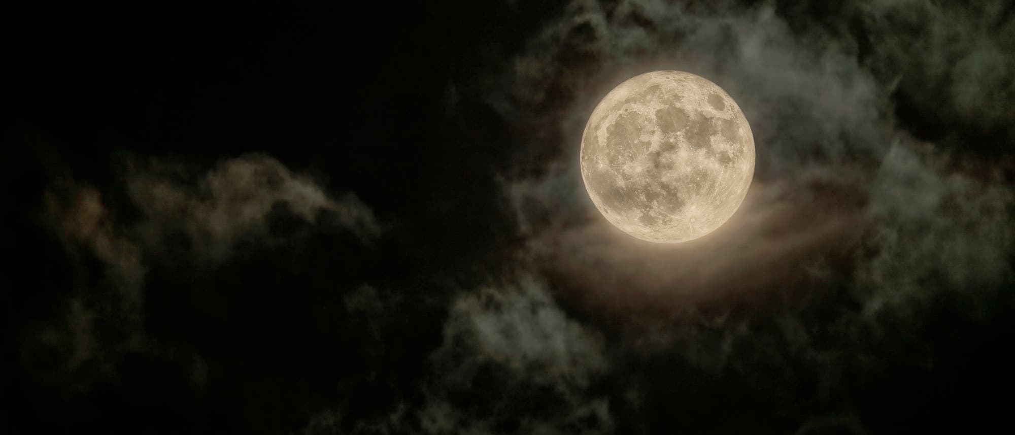 La luna llena brilla intensamente en el cielo nocturno, rodeada de finas y oscuras nubes que le dan un ambiente místico. La superficie de la Luna presenta evidentes cráteres y estructuras propias de su apariencia. El contraste entre la luna brillante y el cielo oscuro enfatiza los detalles de la superficie lunar.