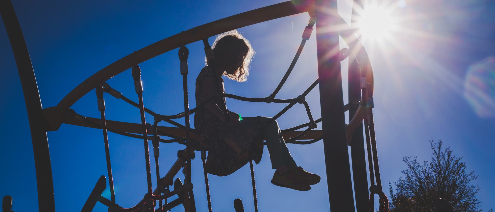 Ein Kind sitzt auf einem Kletterger&uuml;st in einem Spielplatz, umgeben von Seilen und Metallstangen. Die Sonne scheint hell im Hintergrund, wodurch das Kind als Silhouette erscheint. Der Himmel ist klar und blau, und einige B&auml;ume sind im Hintergrund sichtbar.