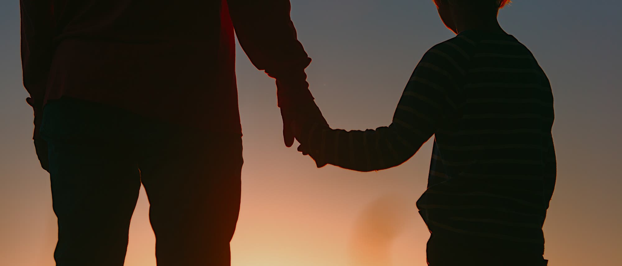 Ein Erwachsener und ein Kind halten sich bei Sonnenuntergang an den Händen. Die Silhouetten sind vor einem farbenfrohen Himmel zu sehen. Die Szene vermittelt ein Gefühl von Geborgenheit und Zusammengehörigkeit. Ein Erwachsener und ein Kind halten sich bei Sonnenuntergang an den Händen. Die Silhouetten sind vor einem farbenfrohen Himmel zu sehen. Die Szene vermittelt ein Gefühl von Geborgenheit und Zusammengehörigkeit.