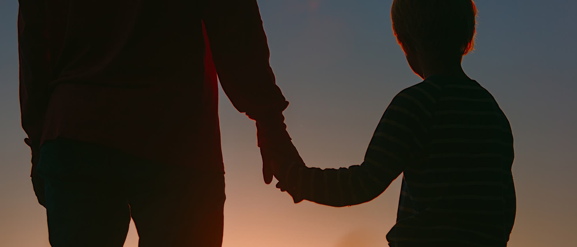 Ein Erwachsener und ein Kind halten sich bei Sonnenuntergang an den Händen. Die Silhouetten sind vor einem farbenfrohen Himmel zu sehen. Die Szene vermittelt ein Gefühl von Geborgenheit und Zusammengehörigkeit. Ein Erwachsener und ein Kind halten sich bei Sonnenuntergang an den Händen. Die Silhouetten sind vor einem farbenfrohen Himmel zu sehen. Die Szene vermittelt ein Gefühl von Geborgenheit und Zusammengehörigkeit.