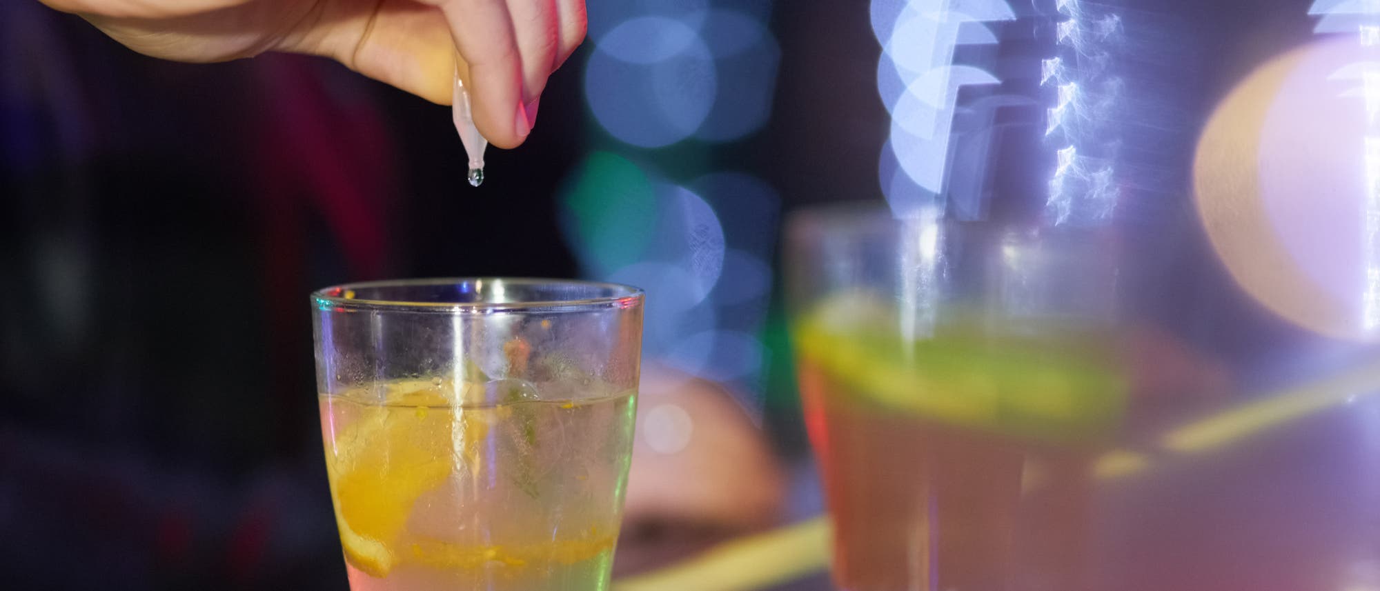 Eine Hand drückt einen Tropfen aus einem kleinen Fläschchen in ein Glas mit einem Getränk, das eine Zitronenscheibe enthält. Im Hintergrund sind verschwommene Lichter zu sehen, die eine lebendige und festliche Atmosphäre schaffen. Das Bild vermittelt den Eindruck einer Bar oder eines Partyumfelds. Eine Hand drückt einen Tropfen aus einem kleinen Fläschchen in ein Glas mit einem Getränk, das eine Zitronenscheibe enthält. Im Hintergrund sind verschwommene Lichter zu sehen, die eine lebendige und festliche Atmosphäre schaffen. Das Bild vermittelt den Eindruck einer Bar oder eines Partyumfelds.