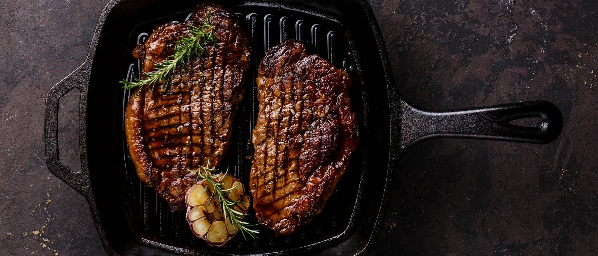 Foto von zwei kross gebratenen Rindersteaks in einer eckigen, gusseisernen Pfanne, garniert mit Rosmarinzweigen