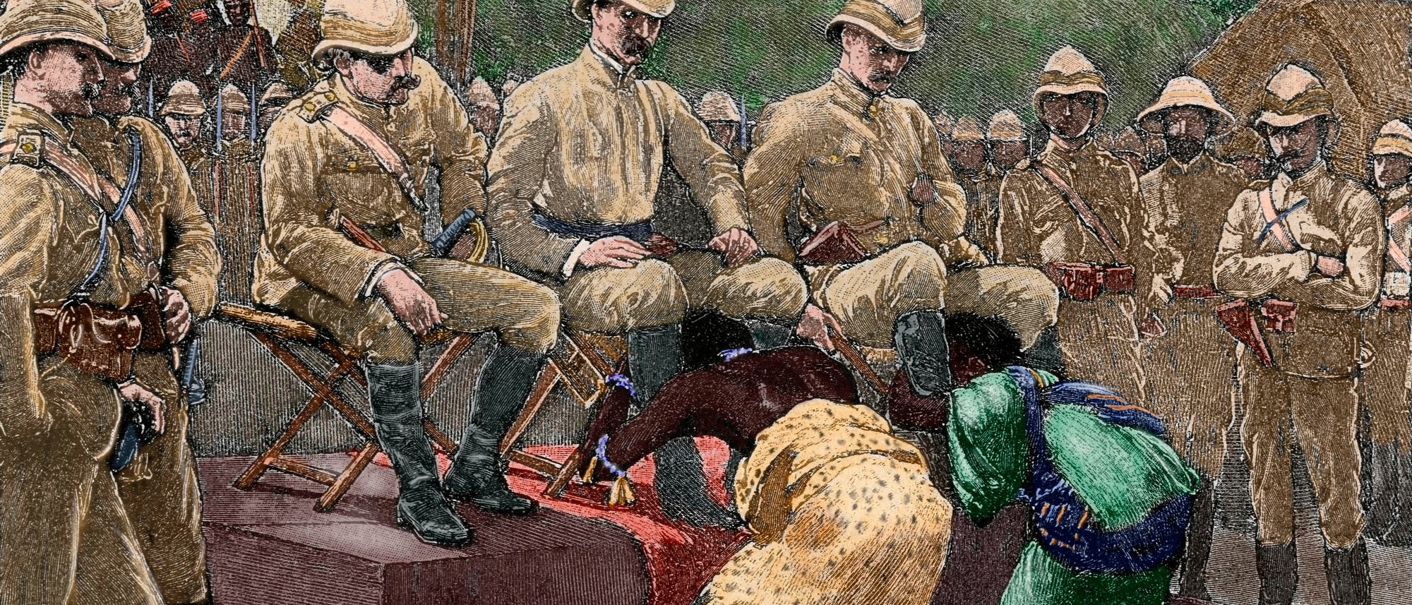 Eine historische Illustration zeigt eine koloniale Szene, in der mehrere uniformierte Männer auf erhöhten Stühlen sitzen. Vor ihnen knien zwei Personen mit gesenkten Köpfen in unterwürfiger Haltung auf einem roten Teppich und umfassen die Beine der Männer. Sie tragen farbenfrohe, traditionell wirkende Kleidung. Im Hintergrund stehen weitere uniformierte Männer in einer Reihe. Die Szene vermittelt ein Gefühl von Hierarchie und Machtungleichheit.