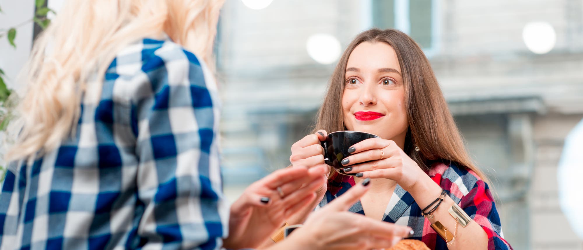 Zwei Personen sitzen in einem Caf&eacute; und unterhalten sich. Eine Person h&auml;lt eine Tasse Kaffee in der Hand und l&auml;chelt, w&auml;hrend die andere gestikuliert. Beide tragen karierte Hemden. Im Hintergrund sind gro&szlig;e Fenster und eine unscharfe Au&szlig;enansicht eines Geb&auml;udes zu sehen. Die Szene wirkt freundlich und entspannt.