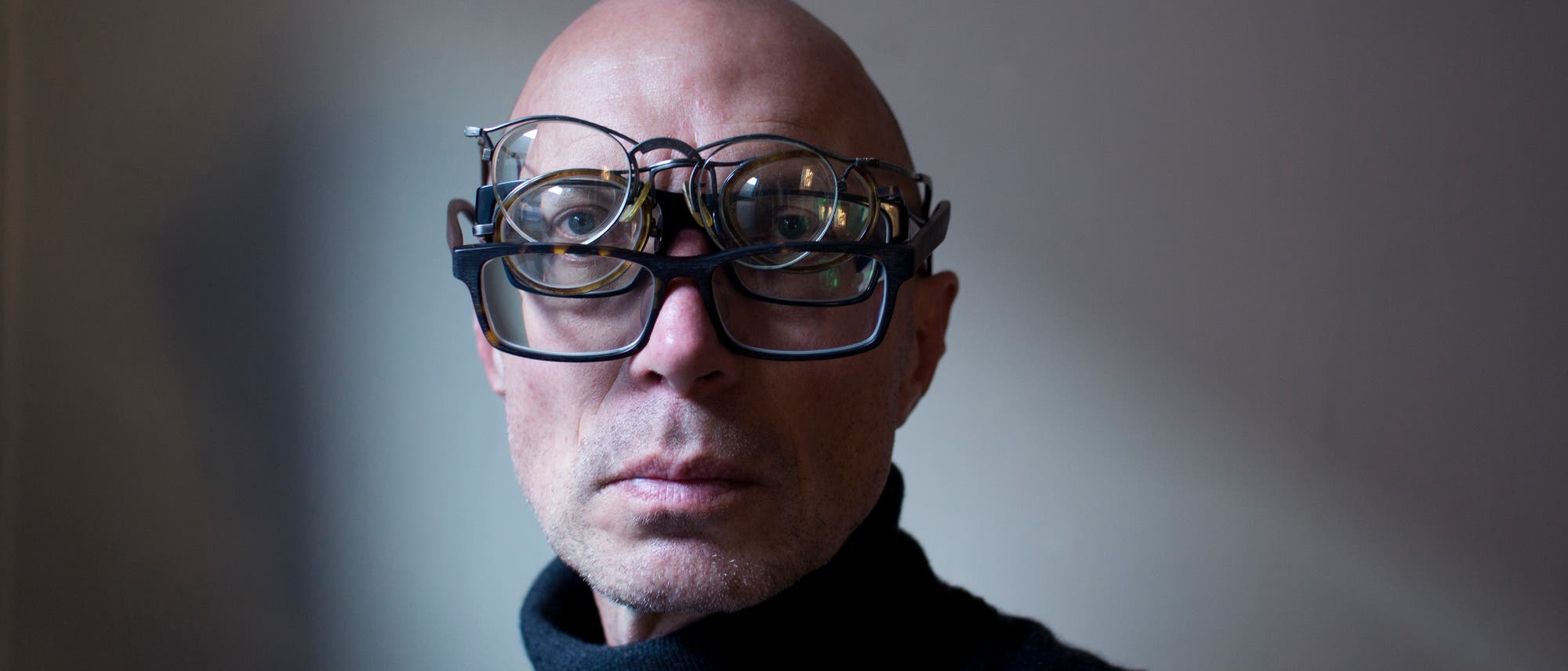 Eine Person mit dunklem Rollkragenpullover tr&auml;gt mehrere Brillen &uuml;bereinander und blickt direkt in die Kamera. Der Hintergrund ist neutral. Die Szene wirkt experimentell.