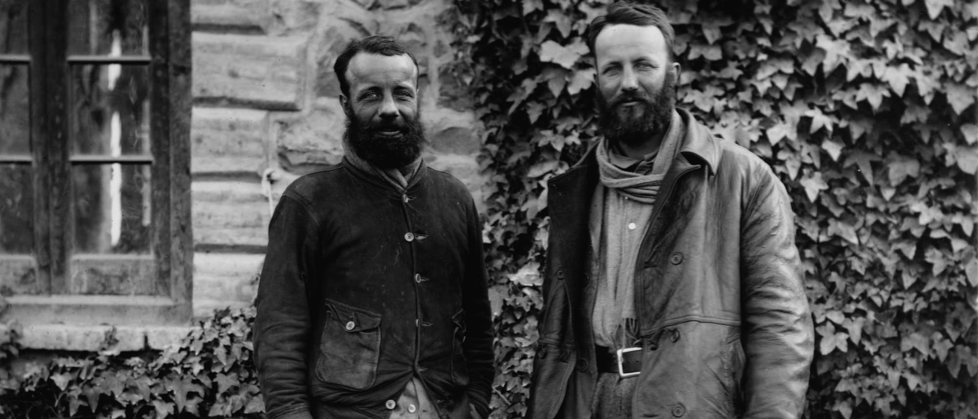 Zwei Männer stehen vor einer mit Efeu bewachsenen Steinwand. Beide tragen wetterfeste Kleidung und haben Bärte. Links im Bild ist ein Fenster zu sehen. Die Szene wirkt historisch und rustikal.