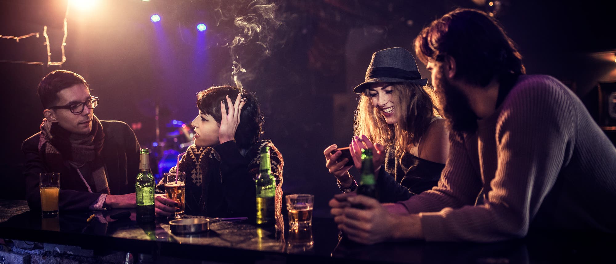 Eine Gruppe von vier Personen sitzt an einer Bar in einem Klub oder einer Lounge. Die Szene ist in gedämpftem Licht gehalten, mit einer warmen, gemütlichen Atmosphäre. Zwei Personen unterhalten sich, während eine andere auf ihr Handy schaut und lächelt. Auf der Theke stehen Getränke, darunter Bierflaschen und Gläser. Im Hintergrund sind bunte Lichter und eine Diskokugel zu sehen, die auf eine entspannte, gesellige Umgebung hinweisen.