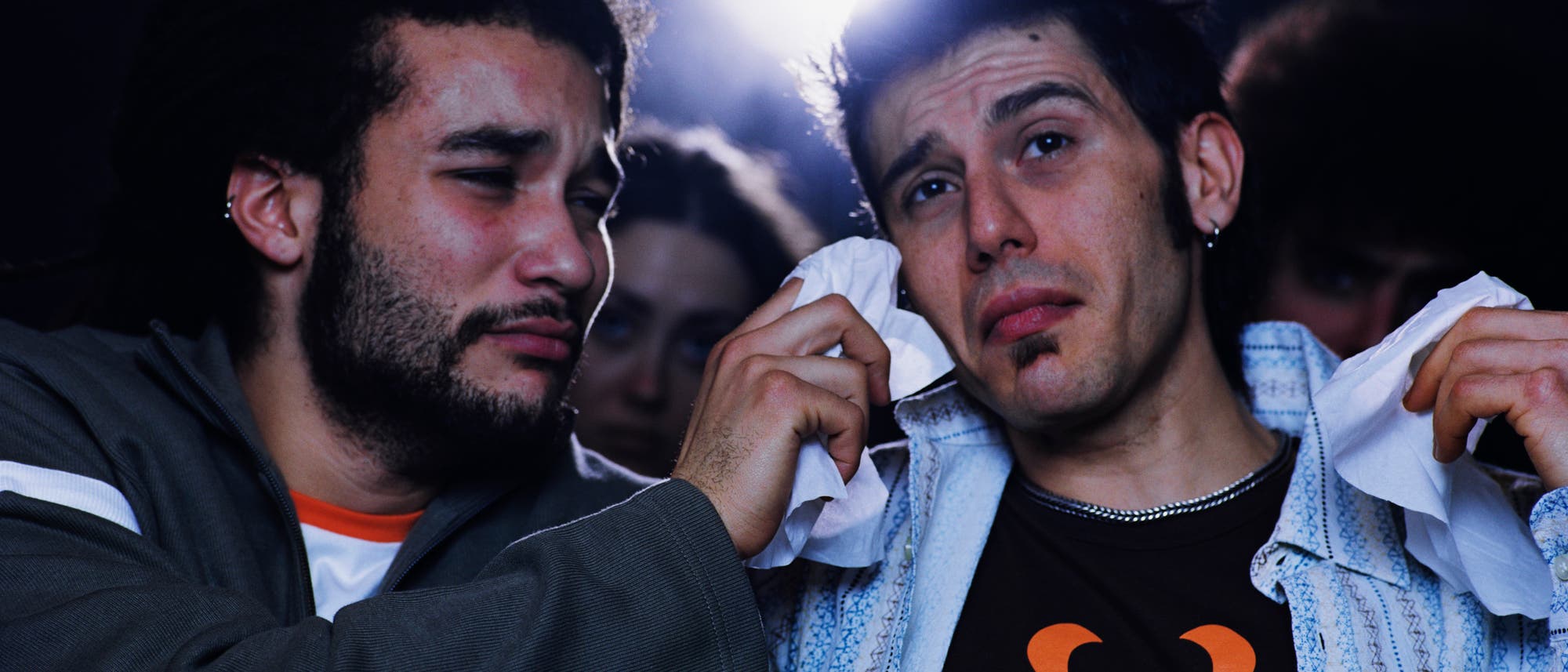 Zwei junge M&auml;nner sitzen nebeneinander im Kino; einer trocknet dem anderen mit einem Taschentuch Tr&auml;nen aus dem Gesicht. Beide wirken emotional bewegt.
