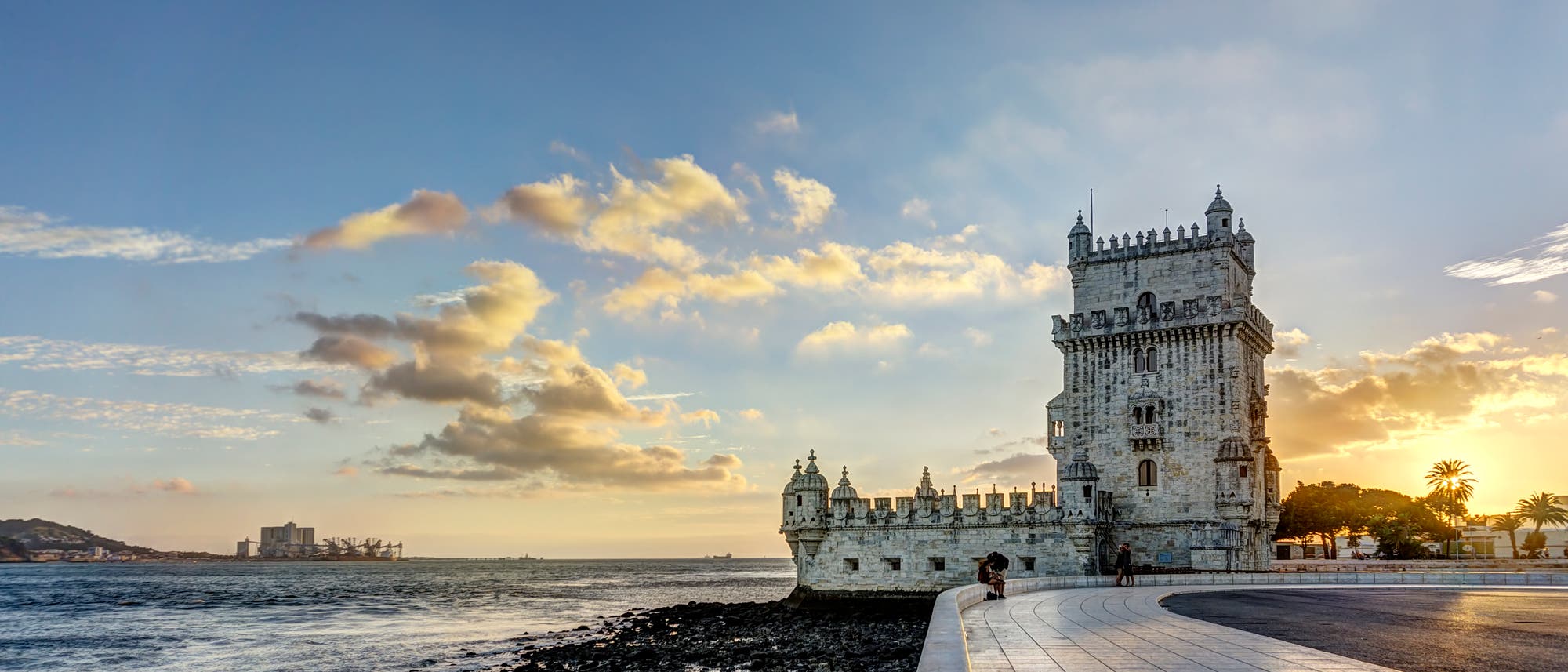 Ein historischer Turm mit Zinnen, Erkern und Türmchen am Ufer eines Flusses bei Sonnenuntergang. Der Himmel ist teils bewölkt, und das Licht der untergehenden Sonne wird auf dem Wasser und den Steinen am Ufer reflektiert. Eine Promenade führt am Wasser entlang, im Hintergrund sind Hügel und Gebäude zu sehen.
