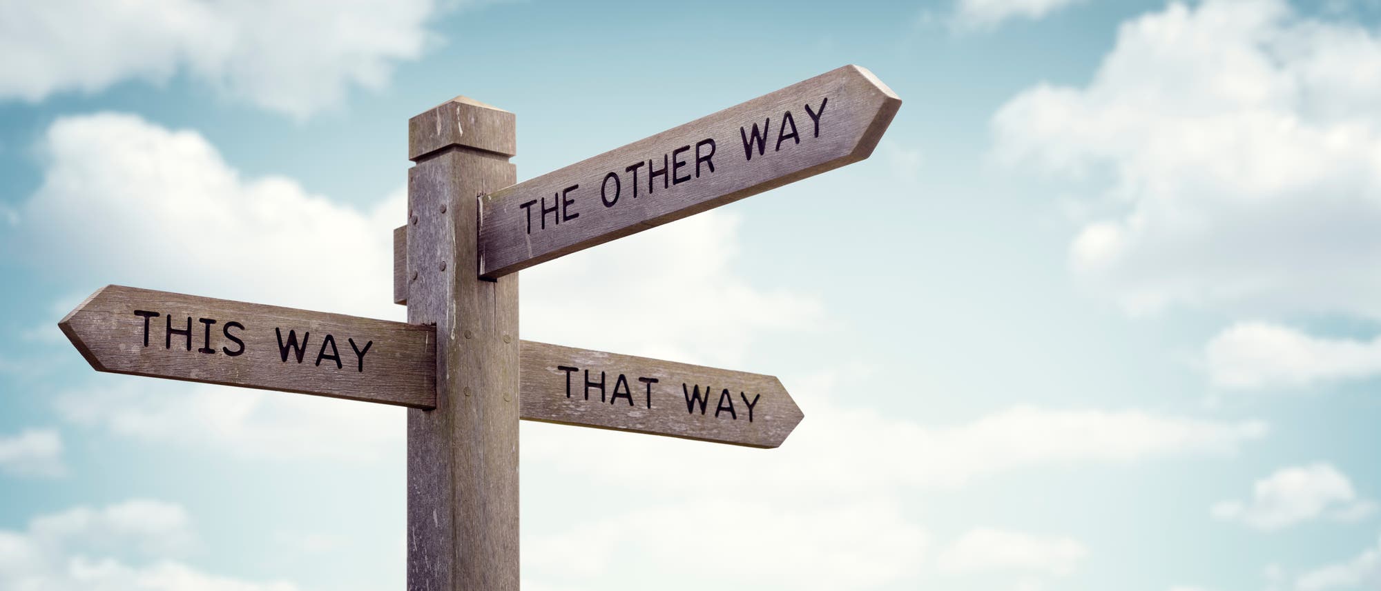 Ein h&ouml;lzerner Wegweiser mit drei Pfeilen zeigt in verschiedene Richtungen. Die Schilder sind mit den Worten &bdquo;This Way&ldquo;, &bdquo;That Way&ldquo; und &bdquo;The Other Way&ldquo; beschriftet. Der Himmel im Hintergrund ist leicht bew&ouml;lkt.