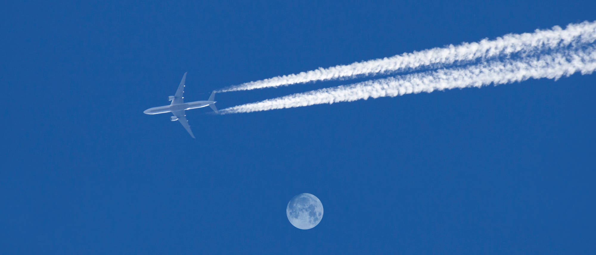 Ein Flugzeug fliegt am klaren blauen Himmel und hinterl&auml;sst zwei Kondensstreifen. Darunter ist der Mond sichtbar.