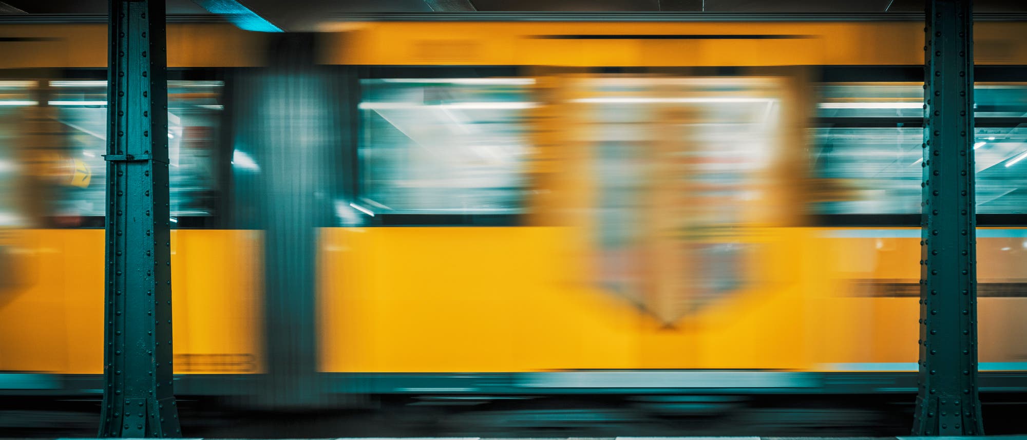 Ein gelber Zug fährt mit hoher Geschwindigkeit durch eine U-Bahn-Station. Die Bewegung des Zuges erzeugt einen verschwommenen Effekt. Im Vordergrund sind die Bahnsteigkanten und metallene Stützpfeiler sichtbar. Die Szene vermittelt Dynamik und urbanes Leben.