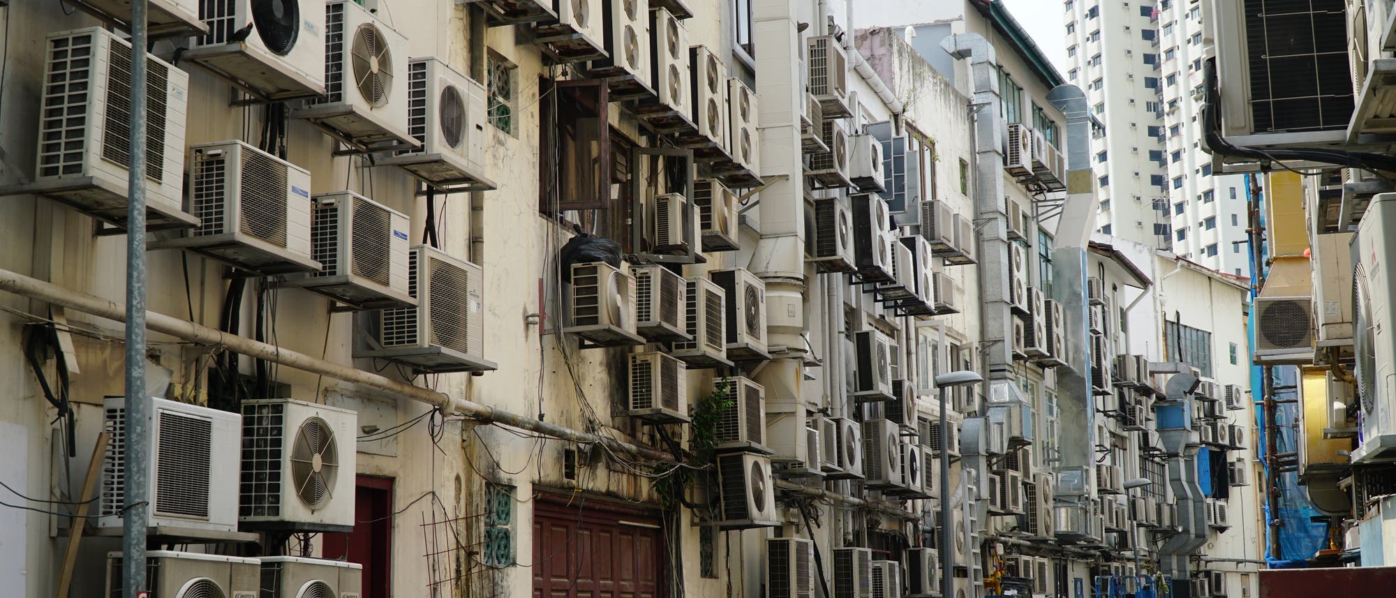 Eine enge Gasse mit zahlreichen Klimaanlagen, die an den Au&szlig;enw&auml;nden der Geb&auml;ude angebracht sind. Die Ger&auml;te sind in mehreren Reihen &uuml;bereinander montiert. Im Vordergrund sind M&uuml;lltonnen und ein Fahrrad zu sehen. Die Szene vermittelt ein Gef&uuml;hl von urbaner Dichte und technischer Infrastruktur.