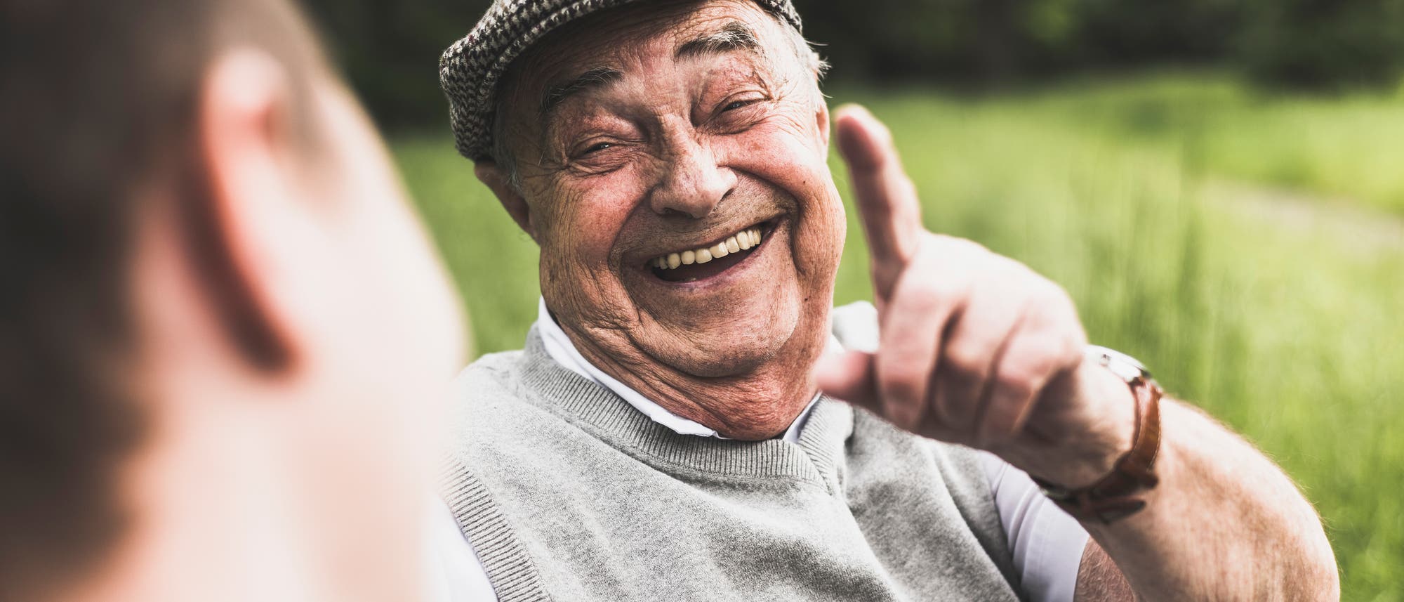 Ein älterer Mann mit einer Schiebermütze lacht herzlich und hebt seinen Finger. Die Szene spielt sich in einem grünen, natürlichen Umfeld ab, was eine entspannte und fröhliche Stimmung vermittelt. Ein älterer Mann mit einer Schiebermütze lacht herzlich und hebt seinen Finger. Die Szene spielt sich in einem grünen, natürlichen Umfeld ab, was eine entspannte und fröhliche Stimmung vermittelt.