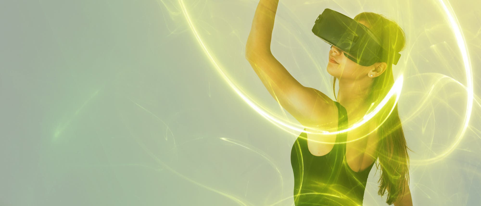 Eine Frau trägt ein Virtual-Reality-Headset und hebt tänzerisch die Hand, wobei sie virtuelle Lichtkreise in Gelb hinterlässt, die sie umgeben. Sie trägt ein schwarzes ärmelloses Oberteil. Die Szene ist in grünlich gelbes Licht getaucht, was eine futuristische Atmosphäre schafft.
