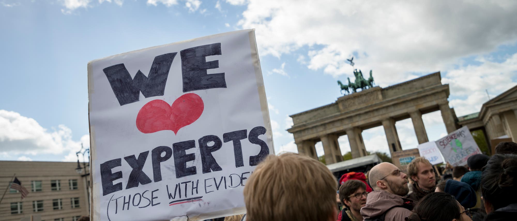 Eine Menschenmenge versammelt sich vor dem Brandenburger Tor. Im Vordergrund hält jemand ein Schild mit der Aufschrift "We ♥ Experts (Those with Evidence)". Der Himmel ist bewölkt, und die Szene vermittelt eine Atmosphäre des Protests bzw. der Unterstützung für wissenschaftliche Expertise. Eine Menschenmenge versammelt sich vor dem Brandenburger Tor. Im Vordergrund hält jemand ein Schild mit der Aufschrift "We ♥ Experts (Those with Evidence)". Der Himmel ist bewölkt, und die Szene vermittelt eine Atmosphäre des Protests bzw. der Unterstützung für wissenschaftliche Expertise.