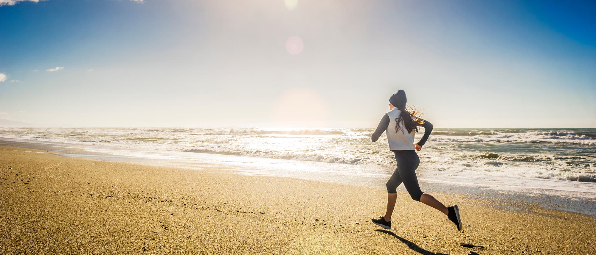 Eine Person joggt am Strand entlang, w&auml;hrend die Sonne am klaren Himmel scheint. Die Wellen des Meeres brechen sanft am Ufer. Die Szene vermittelt ein Gef&uuml;hl von Freiheit und Aktivit&auml;t in der Natur.