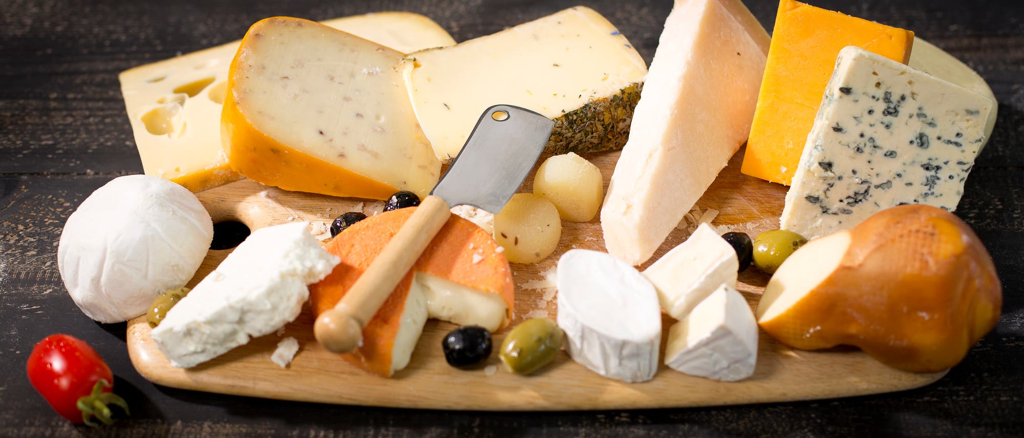 Eine Auswahl verschiedener Käsesorten auf einem Holzbrett, darunter Brie, Blauschimmelkäse, Cheddar und Ziegenkäse. Ein kleines Messer liegt auf dem Brett, umgeben von Oliven, Trauben und einer Kirschtomate. Die Szene ist auf einem dunklen Holztisch arrangiert. Eine Auswahl verschiedener Käsesorten auf einem Holzbrett, darunter Brie, Blauschimmelkäse, Cheddar und Ziegenkäse. Ein kleines Messer liegt auf dem Brett, umgeben von Oliven, Trauben und einer Kirschtomate. Die Szene ist auf einem dunklen Holztisch arrangiert.