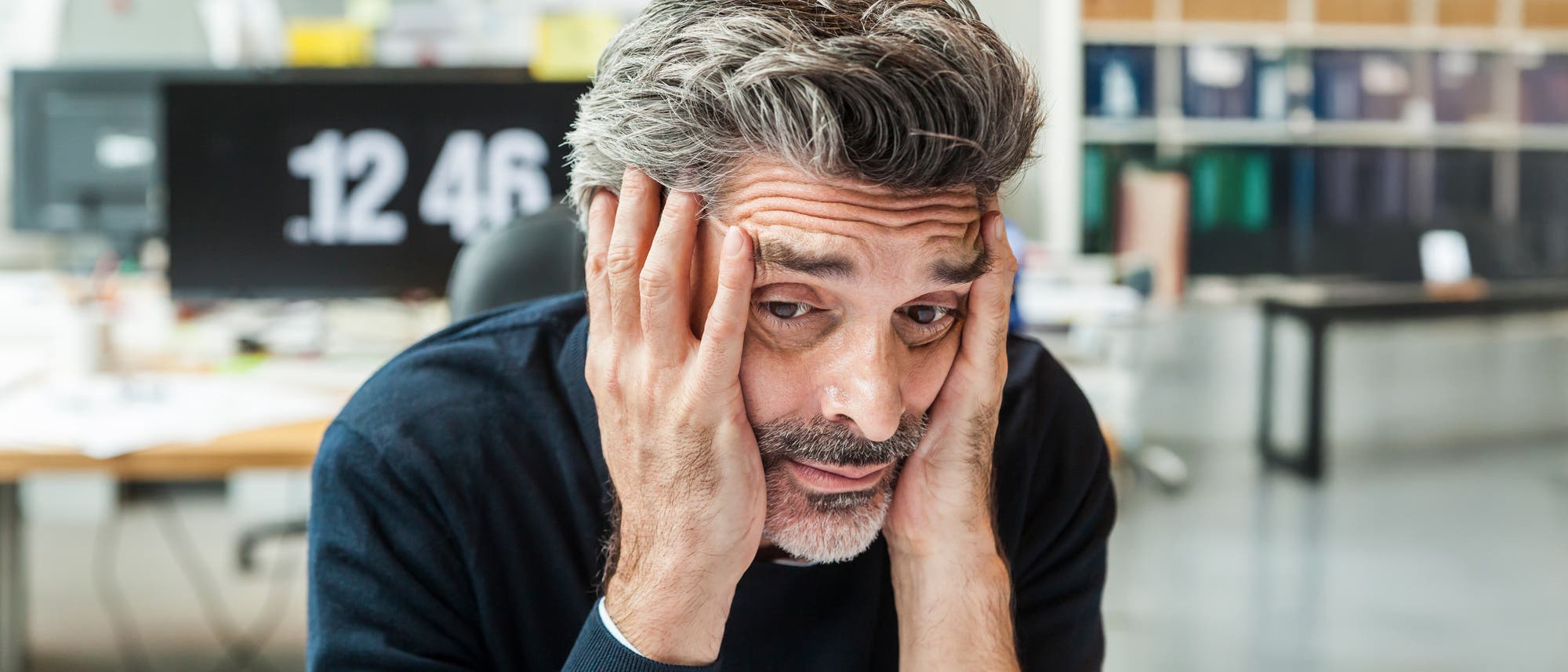 Ein Mann sitzt an einem Schreibtisch und stützt seinen Kopf in die Hände, wirkt nachdenklich oder gestresst. Im Hintergrund ist ein unscharfer Bildschirm mit der Uhrzeit 12:46 zu sehen. Der Raum ist hell beleuchtet und es sind unscharfe Bürogegenstände im Hintergrund erkennbar.