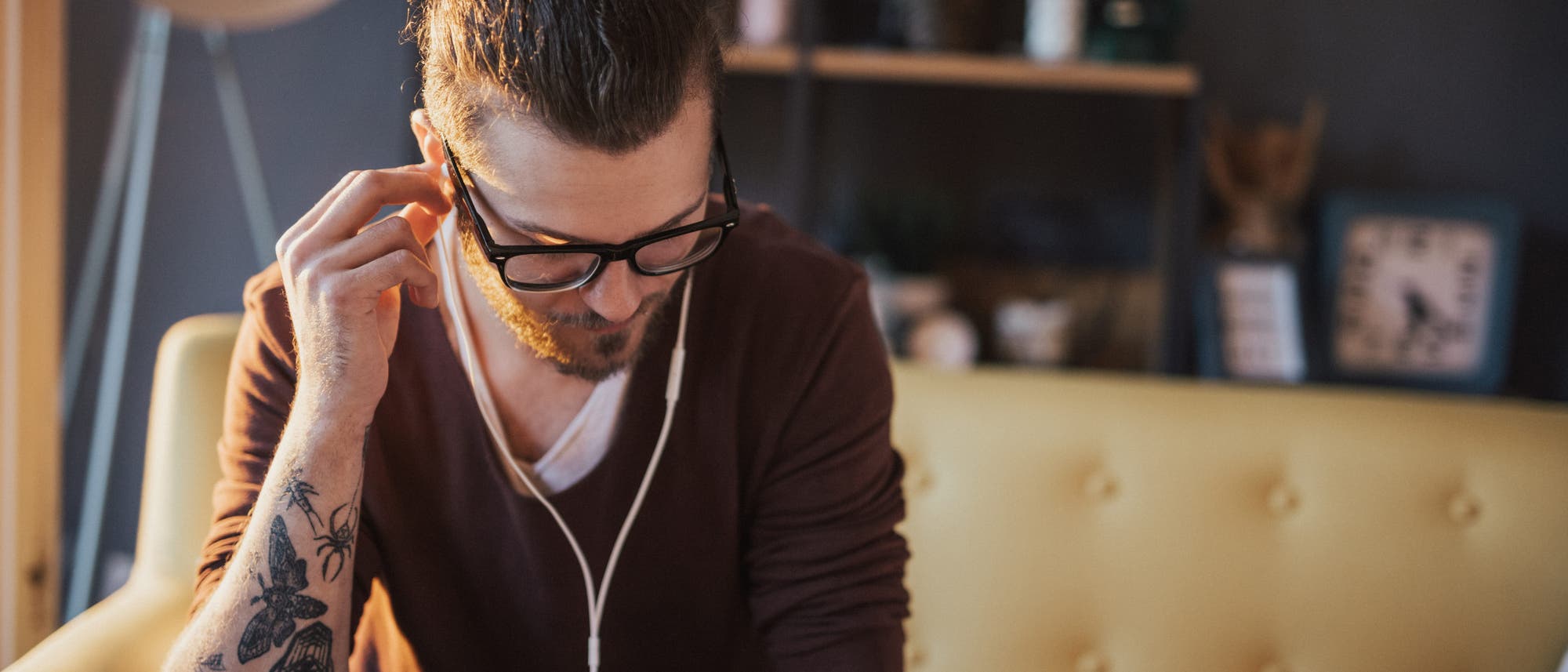 Eine Person sitzt auf einem gelben Sofa und hört etwas über Kopfhörer, die mit einem Smartphone verbunden sind. Im Hintergrund sind ein Globus, eine Uhr und ein Regal zu sehen. Die Person trägt eine Brille und hat ein Tattoo auf dem Arm. Die Szene wirkt entspannt und gemütlich. Eine Person sitzt auf einem gelben Sofa und hört etwas über Kopfhörer, die mit einem Smartphone verbunden sind. Im Hintergrund sind ein Globus, eine Uhr und ein Regal zu sehen. Die Person trägt eine Brille und hat ein Tattoo auf dem Arm. Die Szene wirkt entspannt und gemütlich.