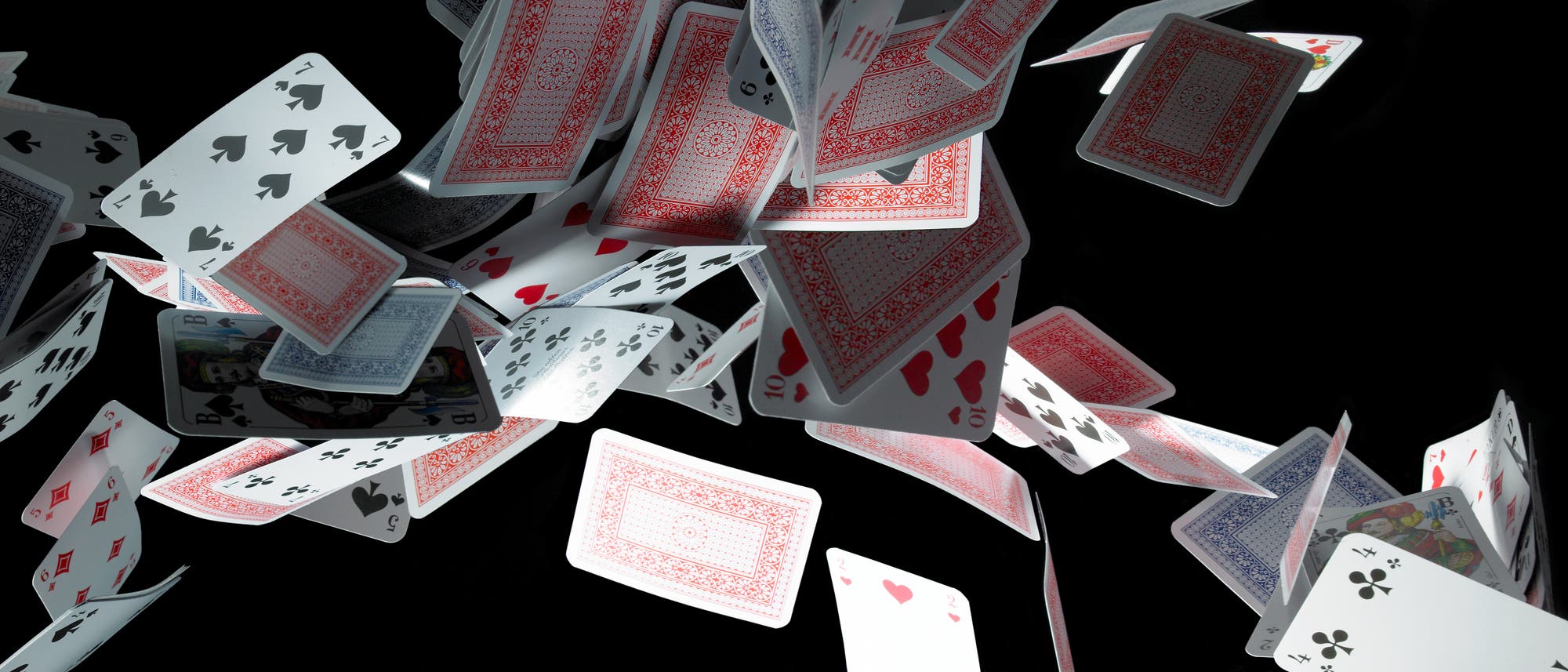 Ein Haufen klassischer Spielkarten fliegt vor einem schwarzen Hintergrund durch die Luft. Die Karten sind in verschiedenen Ausrichtungen sichtbar, mit Vorder- und Rückseiten, die unterschiedliche Farben und Muster zeigen. Einige Karten sind klar erkennbar. Die Szene vermittelt Bewegung und Dynamik.