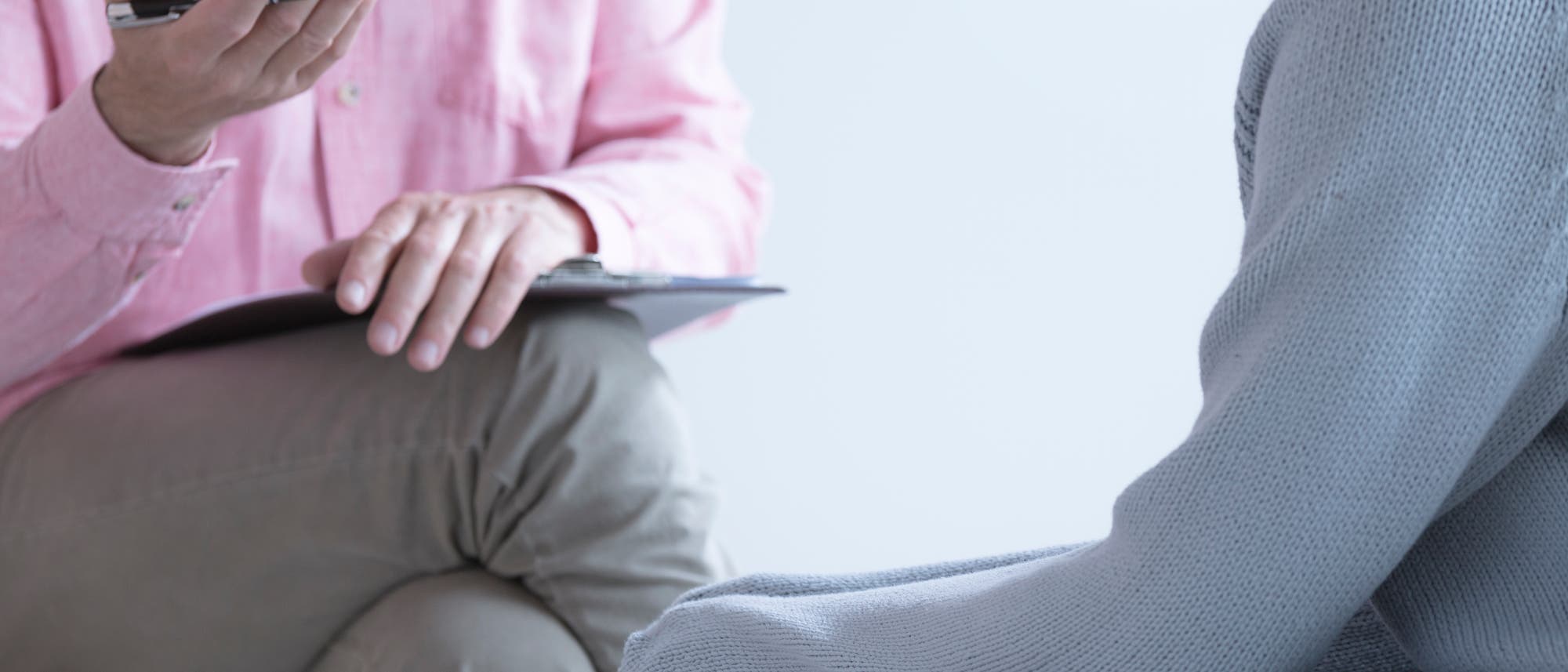 Eine Person in einem rosa Hemd sitzt mit gekreuzten Beinen auf einem Stuhl und hält einen Stift und ein Klemmbrett, während sie mit einer jüngeren Person spricht, die ihre Hände in den Ärmeln eines weiten hellen Pullovers verbirgt. Die Köpfe sind im Ausschnitt nicht gezeigt. Die Szene deutet auf ein Gespräch oder eine Beratungssituation hin.