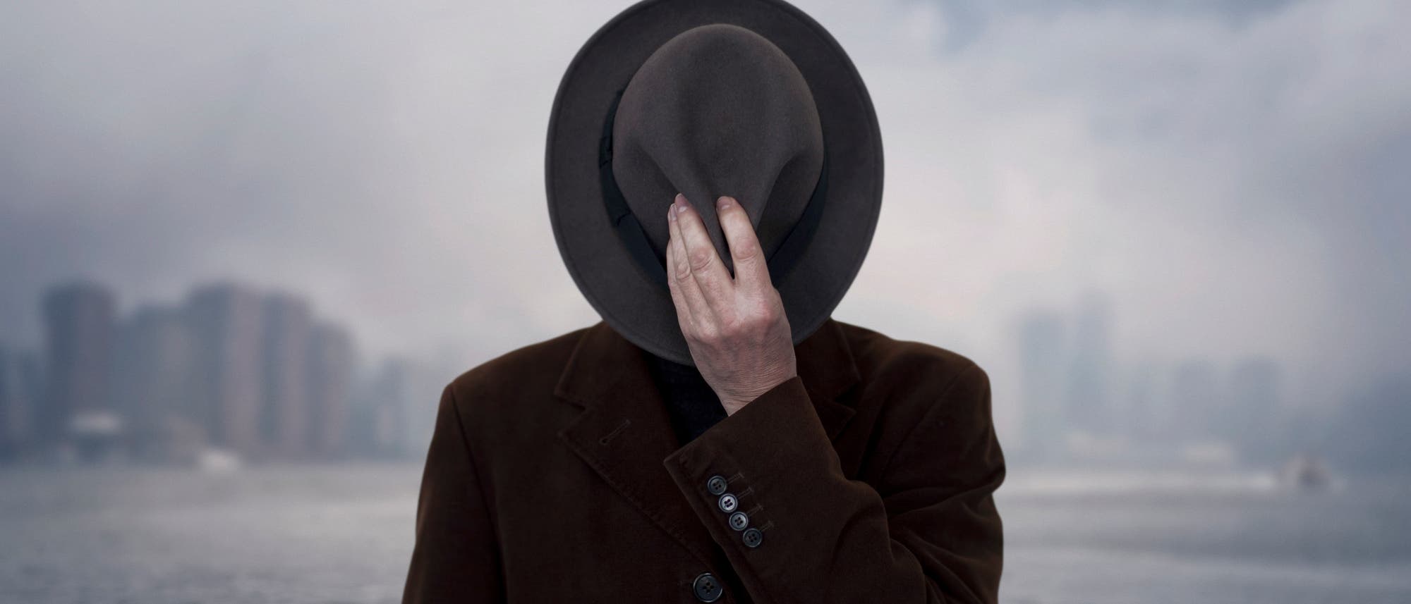 Eine Person steht vor einer verschwommenen Stadtlandschaft am Wasser. Sie trägt einen dunklen Mantel und hält einen Hut, der das Gesicht verdeckt. Der Hintergrund zeigt eine neblige Skyline mit Wolkenkratzern.