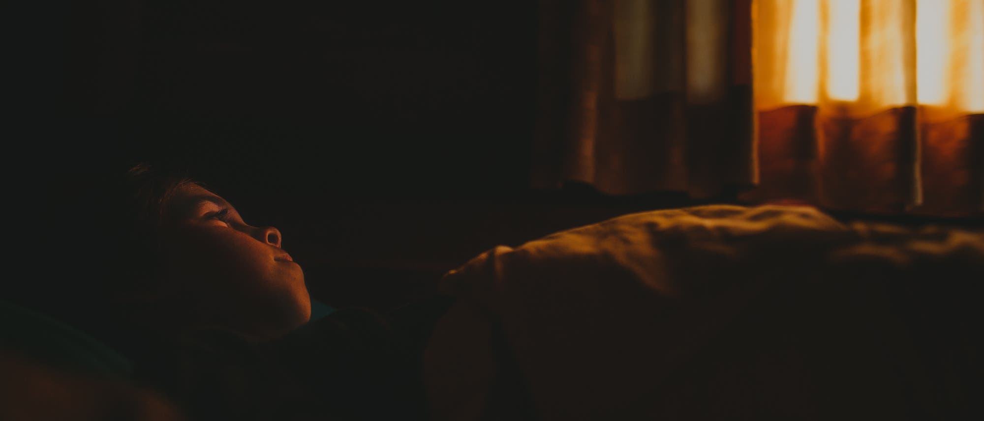 Ein Kind liegt in einem abgedunkelten Raum, während etwas Licht durch ein Fenster mit Vorhängen scheint. Ein Kind liegt in einem abgedunkelten Raum, während etwas Licht durch ein Fenster mit Vorhängen scheint.