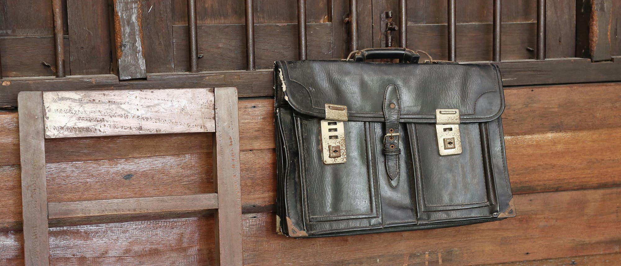 Eine alte, schwarze Ledertasche h&auml;ngt an einer h&ouml;lzernen Wand neben einem einfachen Holzstuhl. Die Tasche hat zwei Metallschl&ouml;sser und zeigt Gebrauchsspuren. Die Szene vermittelt einen rustikalen, nostalgischen Eindruck.