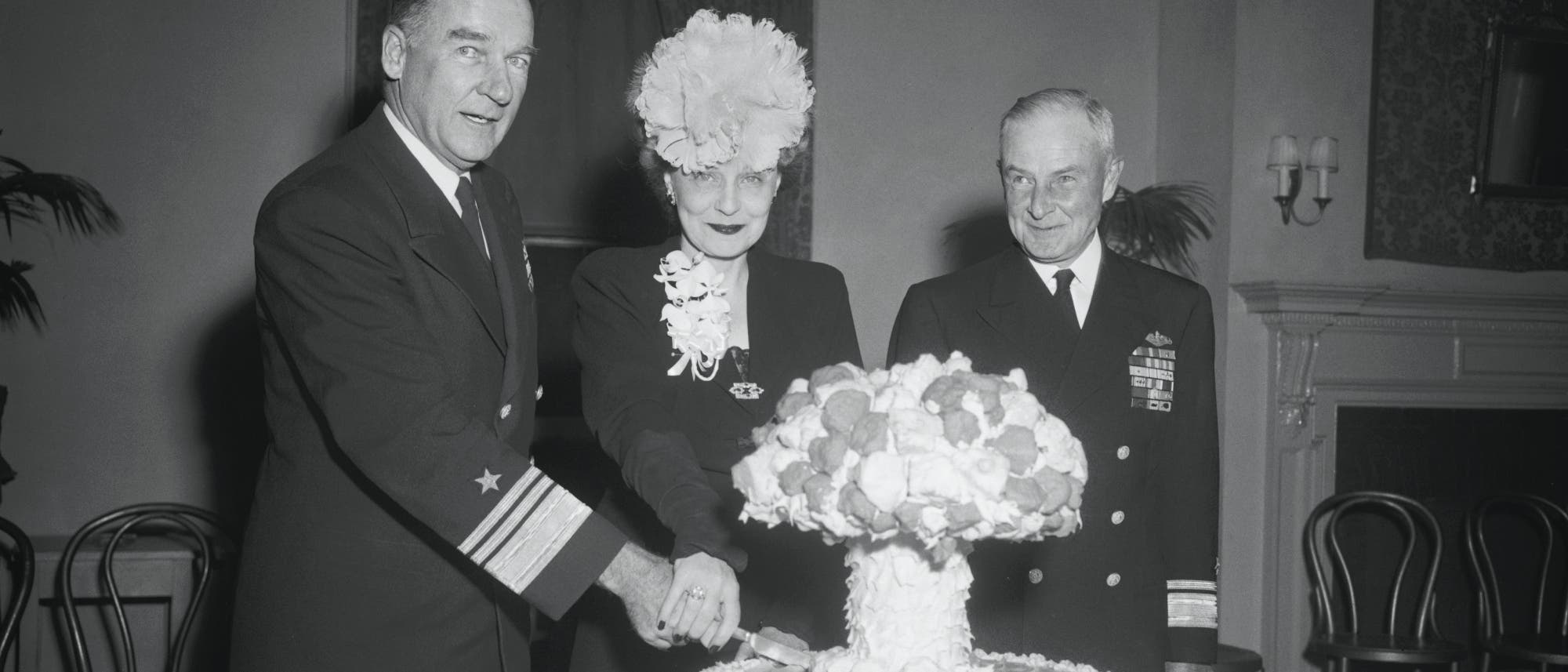 Eine historische Schwarz-Wei&szlig;-Fotografie zeigt drei Personen in formeller Kleidung, die gemeinsam eine Torte in Form einer Pilzwolke anschneiden. Die Torte ist detailliert dekoriert und steht im Mittelpunkt des Bildes. Die Personen l&auml;cheln und wirken feierlich. Im Hintergrund sind M&ouml;bel und eine Wand mit Dekorationen zu sehen.