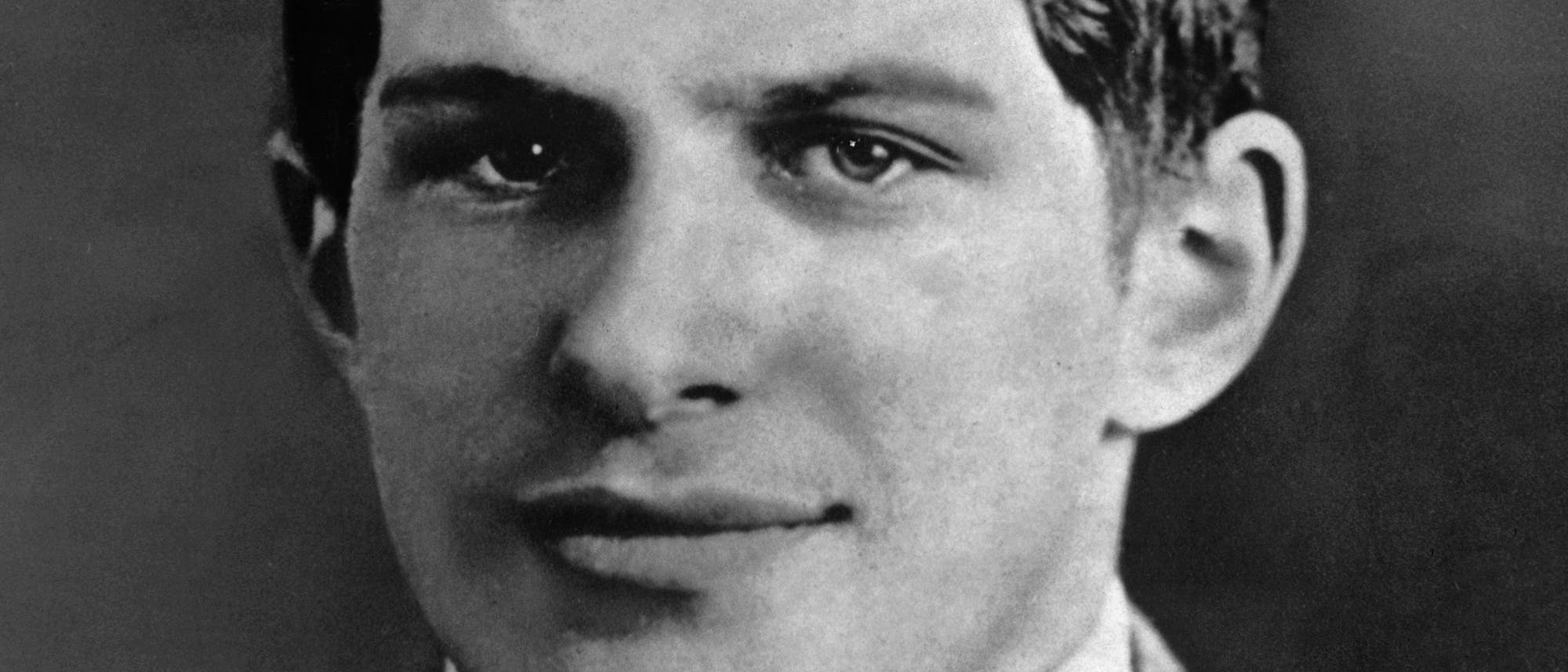 Ein Schwarz-Weiß-Foto zeigt das Gesicht eines Mannes. Der Mann blickt direkt in die Kamera und hat einen neutralen Gesichtsausdruck. Der Hintergrund ist unscharf, wodurch der Fokus auf dem Gesicht des Mannes liegt. 