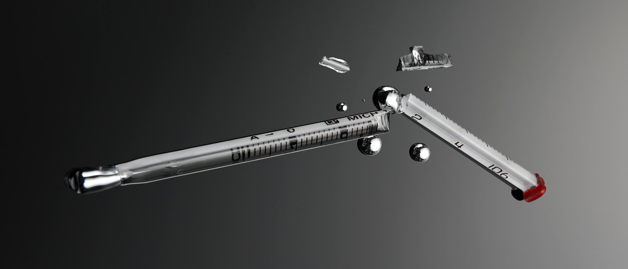 Ein zerbrochenes Glasthermometer liegt auf einer grauen Oberfl&auml;che. Um das Thermometer herum sind kleine Quecksilbertr&ouml;pfchen verteilt. Das Thermometer zeigt eine Skala mit Markierungen und Zahlen. Die Szene vermittelt den Eindruck eines Unfalls oder einer Besch&auml;digung.