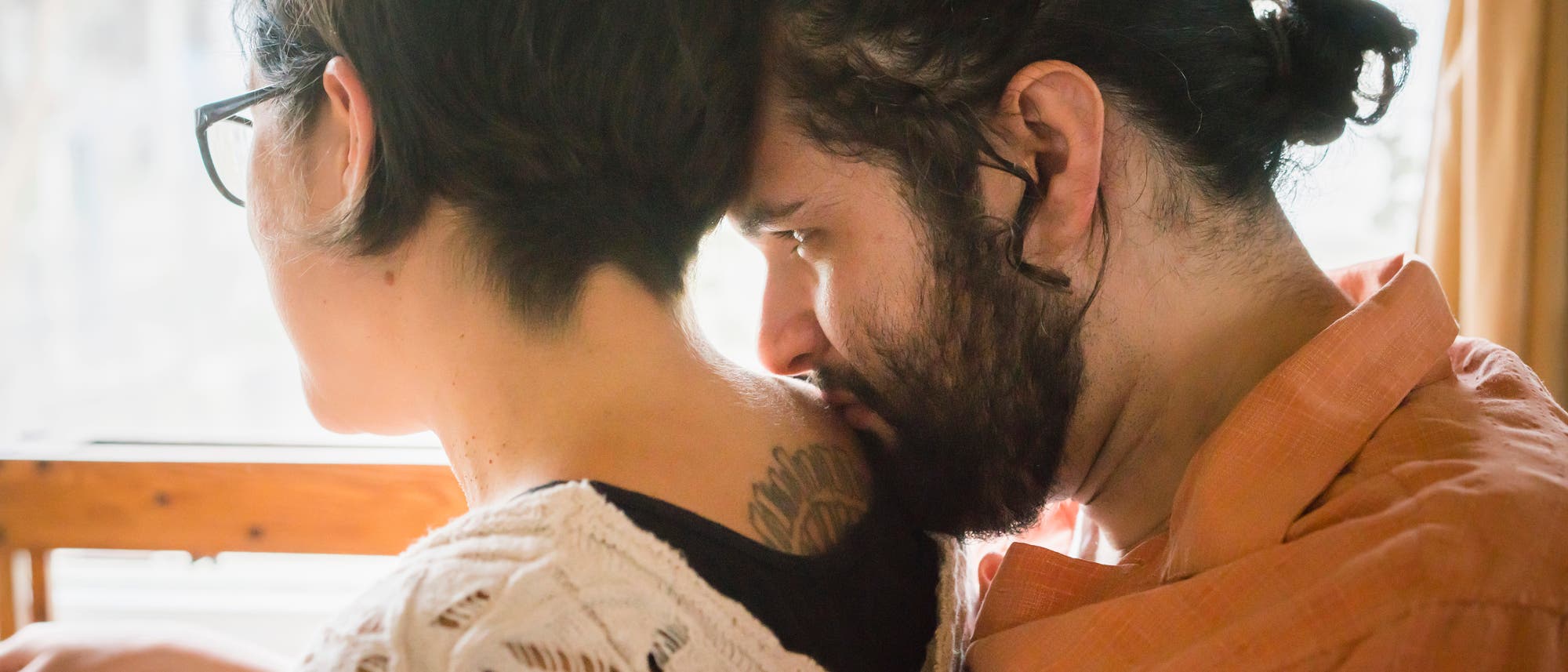 Ein Paar sitzt eng beieinander auf einer Bank am Fenster. Die Person links tr&auml;gt eine Brille und ein gestricktes Oberteil, w&auml;hrend die Person rechts ein orangefarbenes Hemd tr&auml;gt. Beide blicken nach drau&szlig;en, wobei die rechte Person den Kopf sanft auf die Schulter der linken Person legt. Die Szene wirkt ruhig und intim.