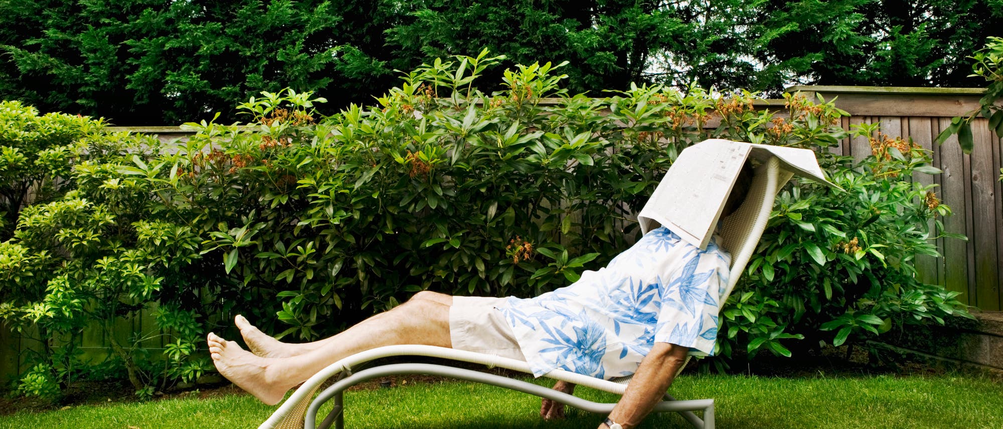 Eine Person liegt entspannt auf einer Sonnenliege im Garten. Sie tr&auml;gt ein Hemd mit blauem Blattmuster und helle Shorts. Eine Zeitung bedeckt ihr Gesicht, w&auml;hrend sie sich ausruht. Im Hintergrund sind gr&uuml;ne B&uuml;sche und eine Holzzaun zu sehen. Die Szene vermittelt eine ruhige und entspannte Atmosph&auml;re.
